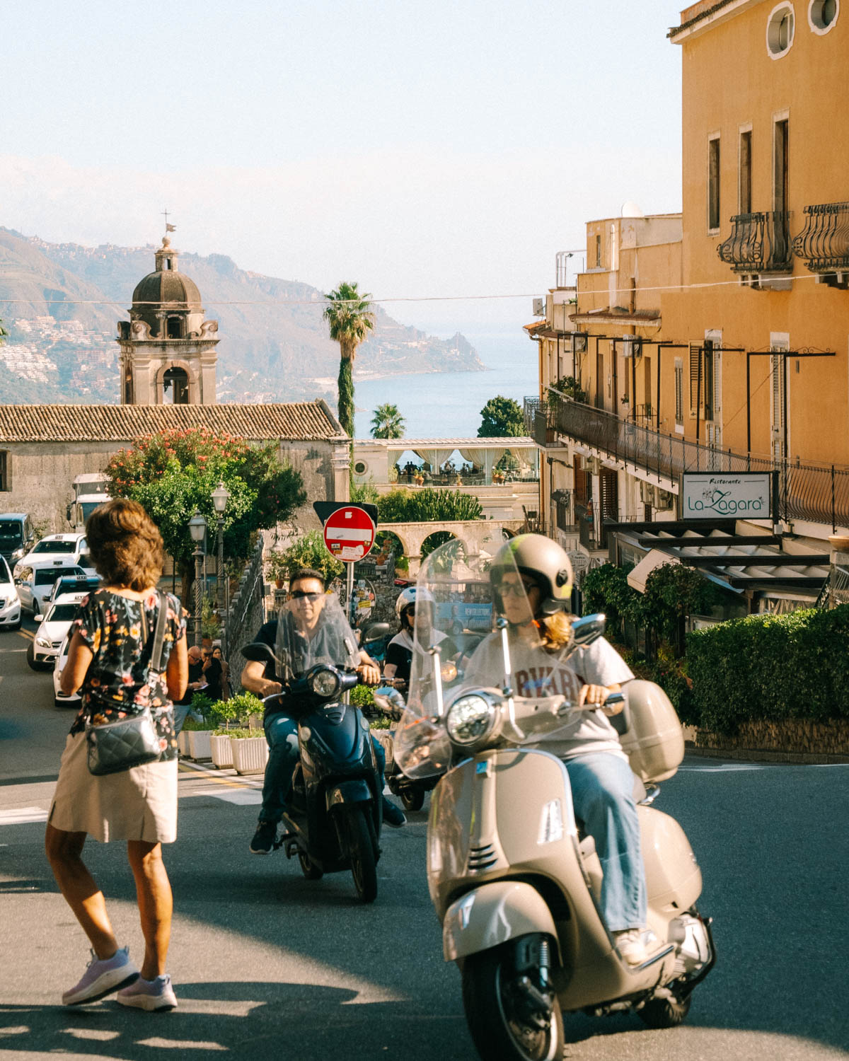 Street scene in Taormina, Sicily, with scooters and pedestrians in the foreground, colorful buildings, and a view of the coastline and mountains in the background