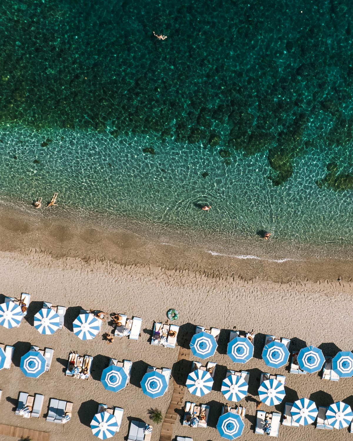 Aerial view of Taormina beach with blue umbrellas and turquoise water.
