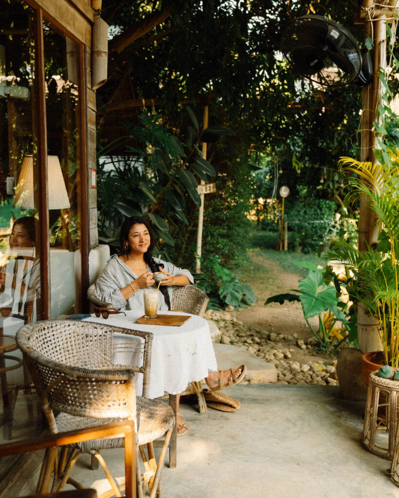 A woman relaxing with an iced coffee on the outdoor terrace of Din Cafe. She is seated at a cozy wicker table, surrounded by tropical plants and bathed in warm golden light, enjoying the cafe’s tranquil ambiance