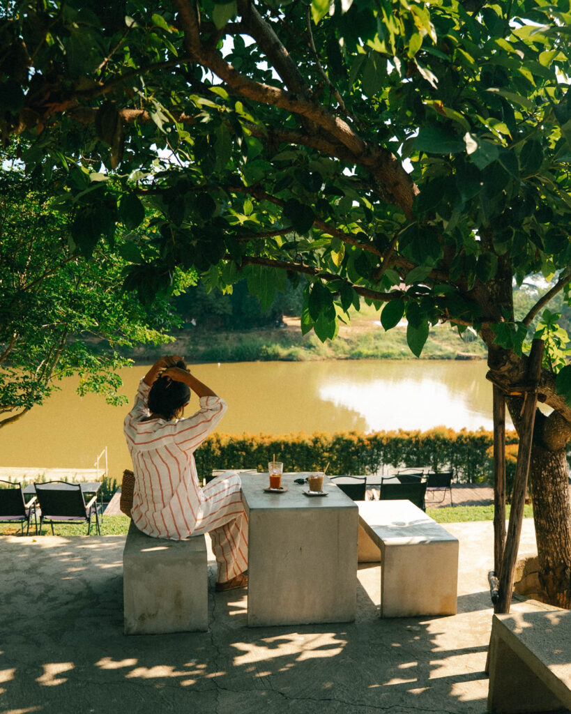 A woman in a striped outfit sitting at a concrete table under the shade of a tree at The Baristro x Ping River. She enjoys her drinks while gazing at the peaceful riverside scenery, surrounded by dappled sunlight and lush foliage