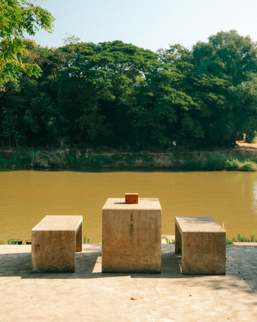 A simple concrete table and benches directly facing the Ping River, offering a tranquil setting for guests at The Baristro. The golden sunlight casts a warm glow over the water, creating a peaceful atmosphere ideal for unwinding