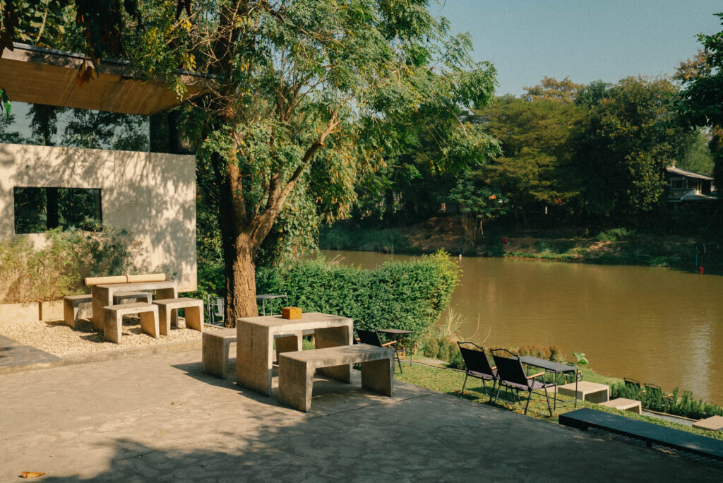 The outdoor seating area of The Baristro x Ping River, featuring minimalist concrete tables and benches overlooking the calm waters. Surrounded by lush greenery and modern architecture, this serene riverside setting is perfect for a relaxed coffee break