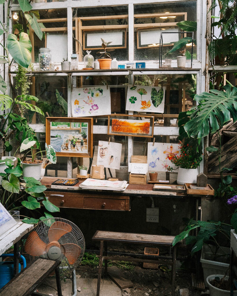 A creative artist's corner at Baankangwat artist market chiang mai, featuring a rustic wooden desk adorned with botanical watercolor paintings, sketches, and art supplies. The surrounding glass windows let in natural light, while potted plants and vines add to the lush, organic feel of the space.