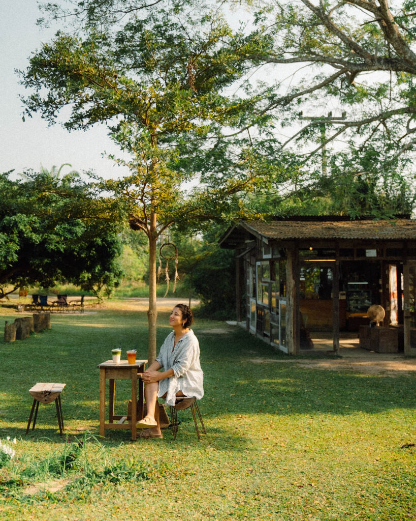 A woman enjoying a peaceful moment at Far Away Cafe Chiang Mai Thailand, sitting at a wooden table under a tree in the countryside. The rustic cafe building in the background, lush greenery, and golden sunlight create a serene and inviting atmosphere