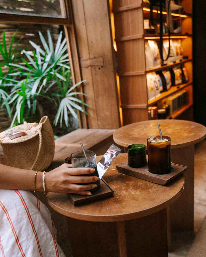 A hand reaching for an iced charcoal latte at Graph Cafe in Chiang Mai, served in a dark minimalist glass. The rustic wooden tables and warm ambient lighting create a cozy atmosphere, with a woven bag and lush green plants in the background