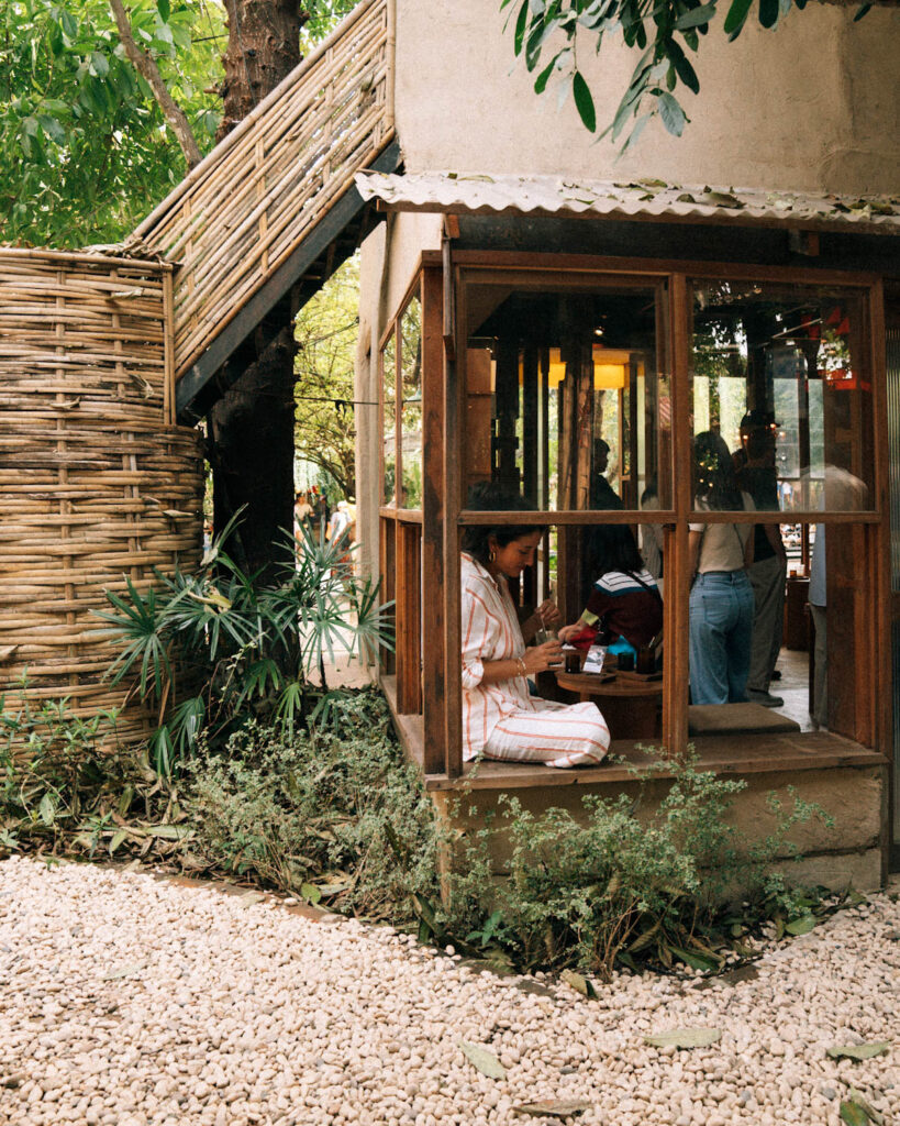 A woman in a striped dress sitting by a window nook at Graph Cafe, enjoying her coffee in a peaceful setting. The earthy tones of the cafe’s architecture, woven bamboo fencing, and greenery give it a charming, natural vibe