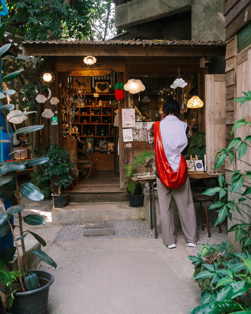 A customer browsing a small artisan shop at Baankangwat artist market Chiang Mai, filled with handcrafted goods, trinkets, and warm glowing lights. The wooden hut-style storefront, surrounded by plants and unique hanging lamps, enhances the cozy, hidden-gem feel of the cafe