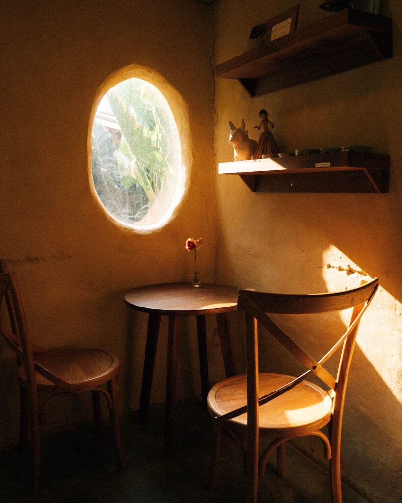 A warmly lit, intimate corner inside Din Cafe featuring a small round wooden table and two chairs. Sunlight filters through a uniquely shaped oval window, casting soft shadows on the textured earth-toned walls, with wooden shelves displaying small decorative items