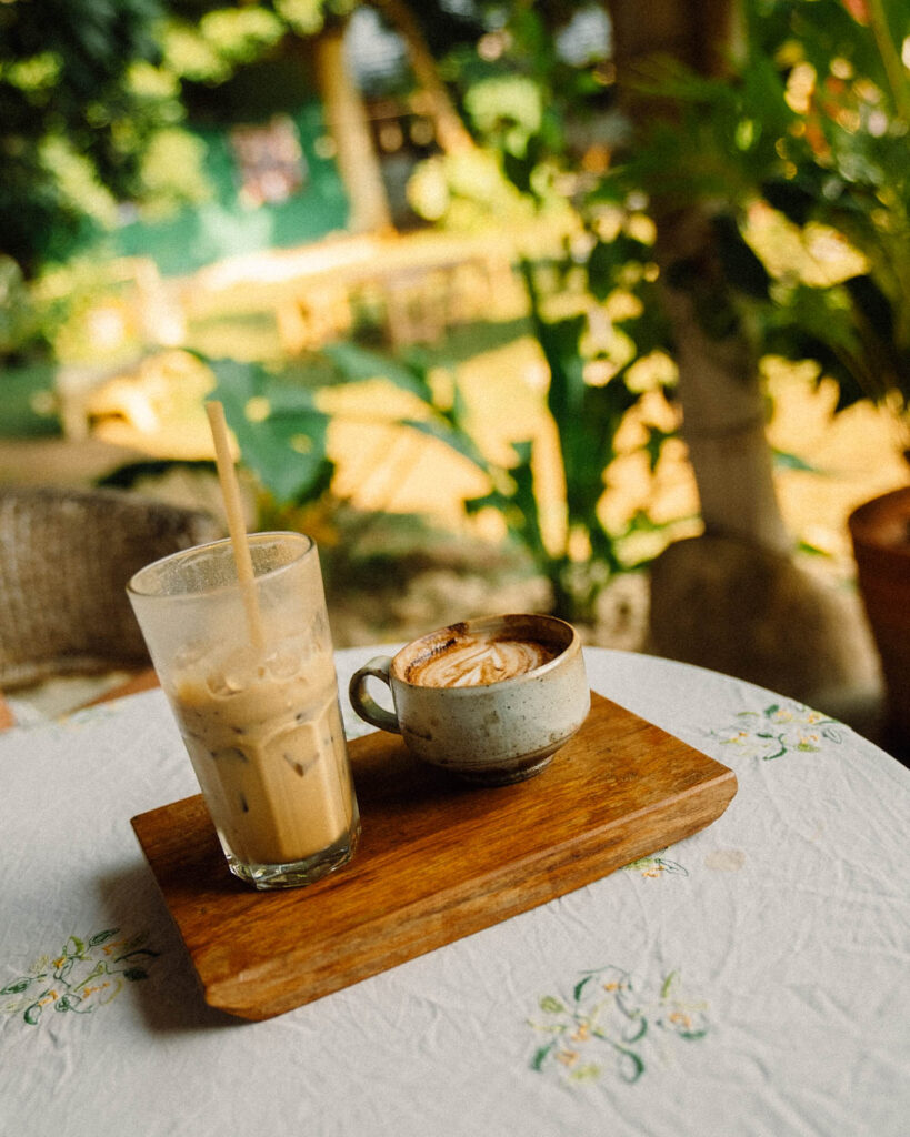 A wooden tray with a handcrafted ceramic cup of cappuccino and a tall glass of iced coffee, set on a white floral tablecloth. The blurred green garden in the background enhances the serene and natural atmosphere of Din Cafe