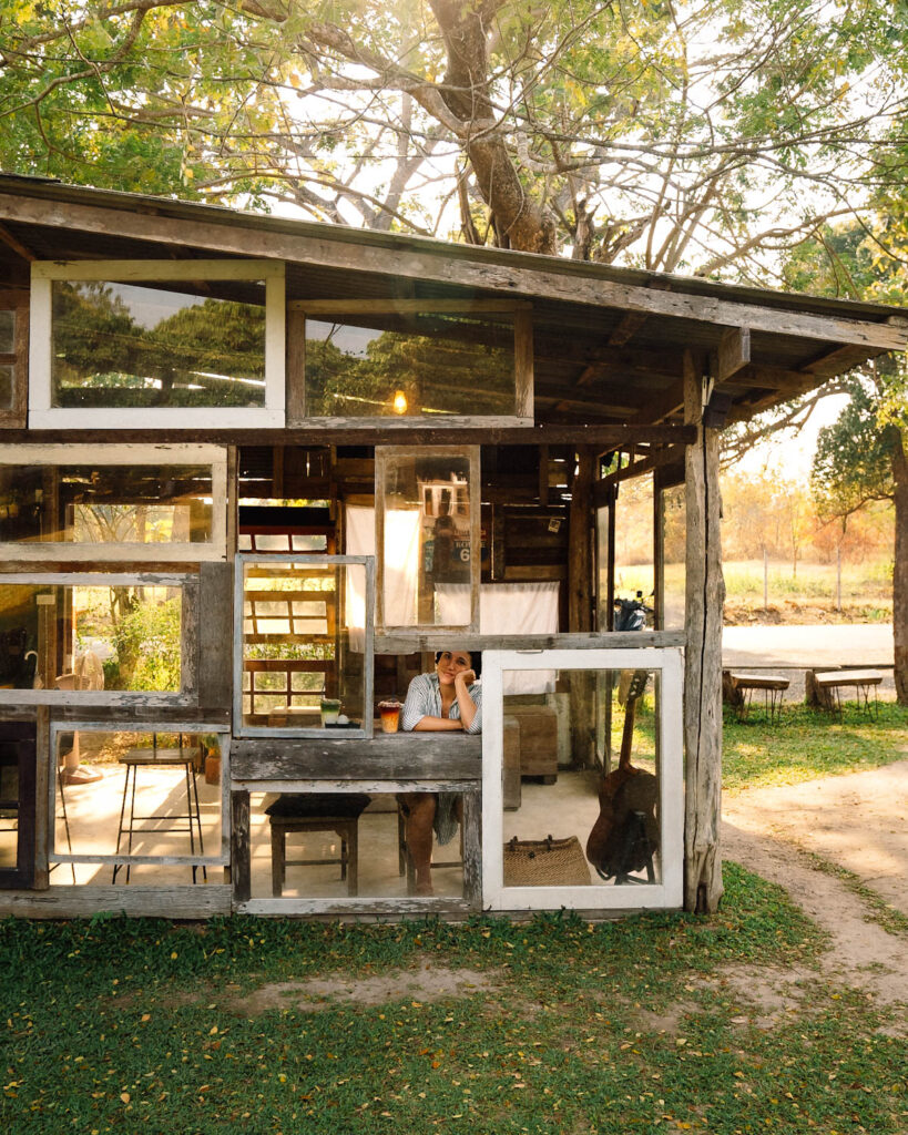 A unique glass-paneled wooden cafe structure at Far Away Cafe, blending seamlessly with nature. A woman sits inside by an open window, gazing into the distance, surrounded by a peaceful rural landscape bathed in golden-hour light