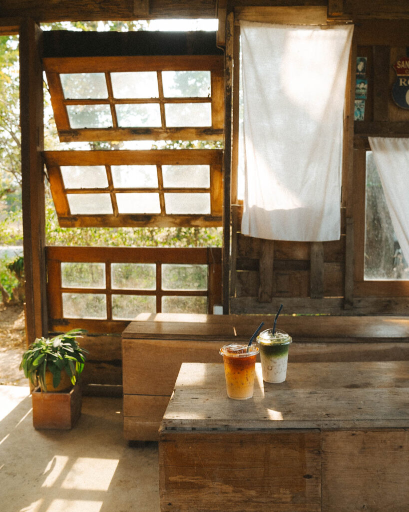 Two iced drinks, a layered Thai tea and a matcha latte, placed on a rustic wooden table inside Far Away Cafe in Chiang Mai. Sunlight filters through vintage wooden windows and soft linen curtains, adding to the cafe’s warm and cozy ambiance