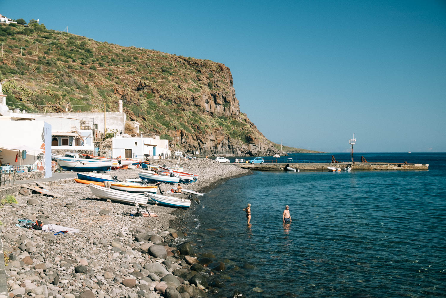 white washed houses and Fishing boats line a stony beach with 2 bathers swimming on Filicudi island Sicily