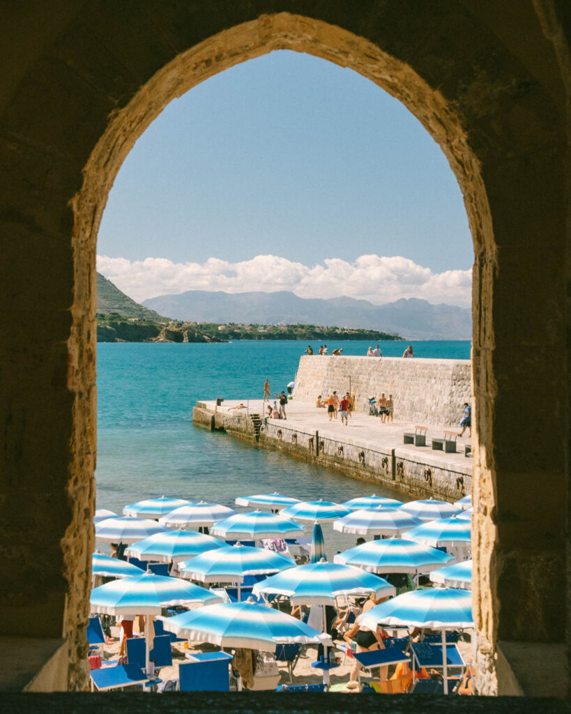 an stone arch frames a summer beach scene of blue umbrellas and turquoise waters in cefalu sicly
