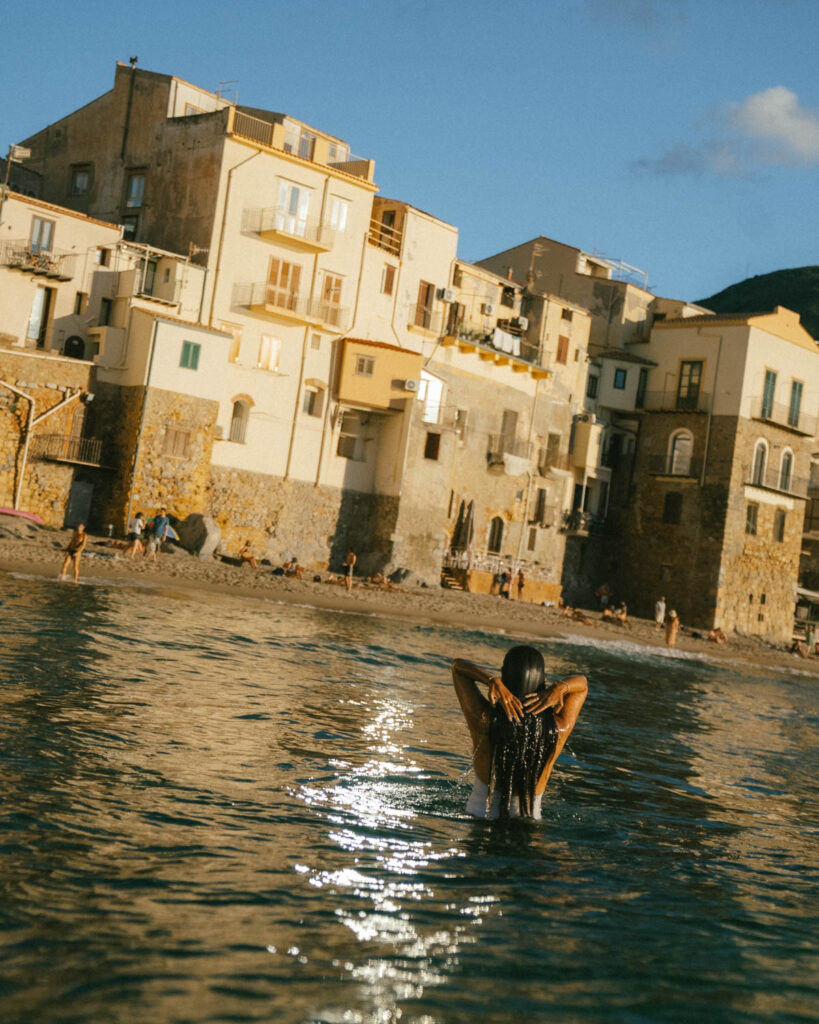 a woman swims at sunset at Spiaggia del Porto Vecchio cefalu with the white washed building lit with golden afternoon light