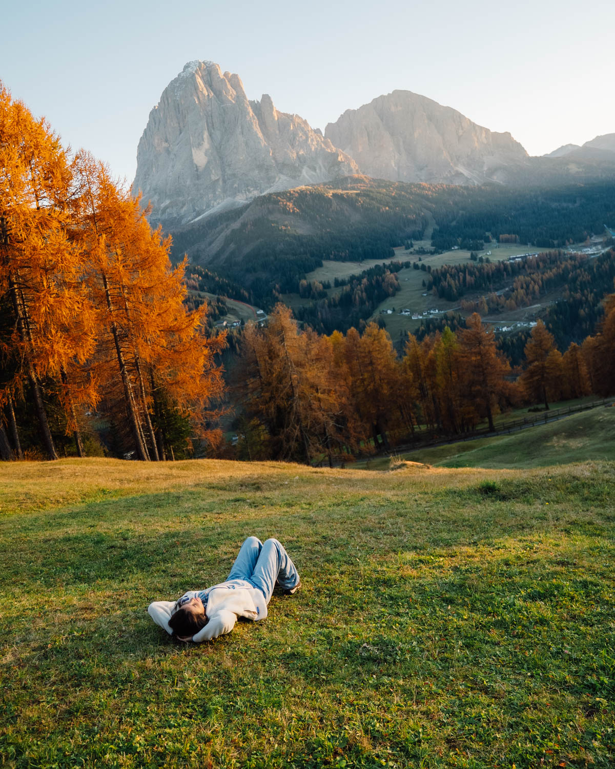 A person lies on a grassy hill, arms behind their head, gazing at the golden larches and towering peaks of the Dolomites. The warm autumn sunlight casts a dreamy glow over the landscape.