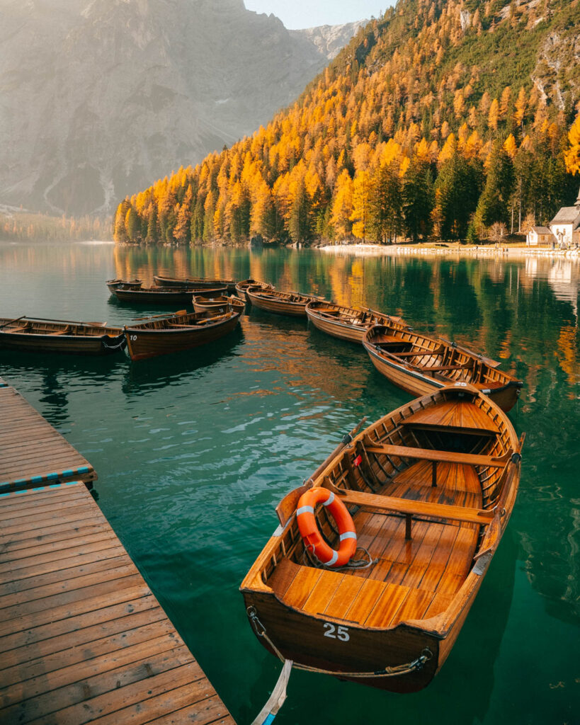 A row of wooden rowboats floats on the emerald waters of a tranquil alpine lake. The shoreline is lined with golden autumn trees, and the towering Dolomites create a breathtaking backdrop.