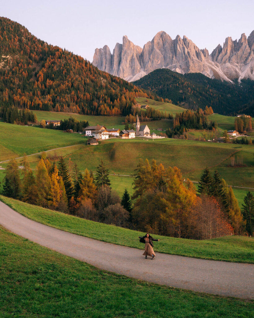 A person twirls on a winding path through a valley in Santa Maddalena with rolling green hills, colorful autumn trees, and a picturesque village at the foot of the Dolomites. The jagged mountain peaks tower in the distance.