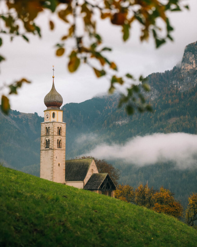 historic St. Valentin church with a bell tower sits on a sloping green hill. The scene is framed by blurred autumn leaves in the foreground, with misty mountains in the background.