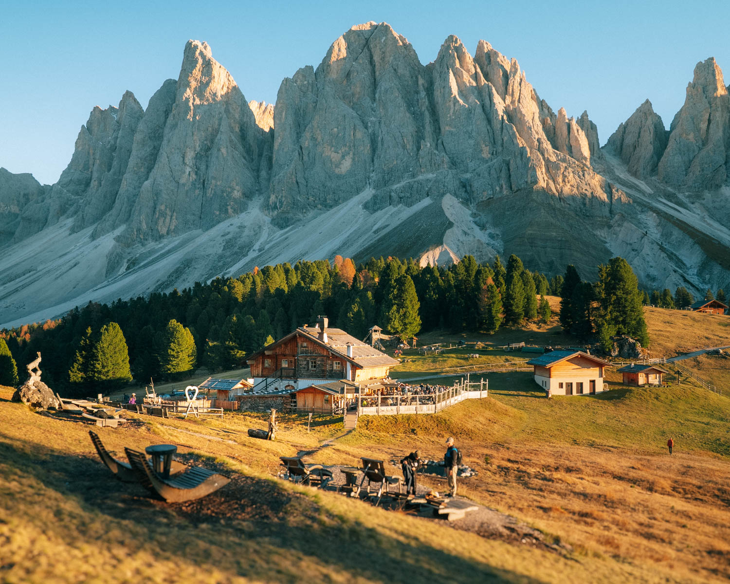 Rifugio delle Odle traditional alpine hut nestled at the base of the jagged Dolomites peaks, surrounded by golden-hued trees and wooden chalets. Sun loungers face the dramatic mountain backdrop, bathed in golden light.