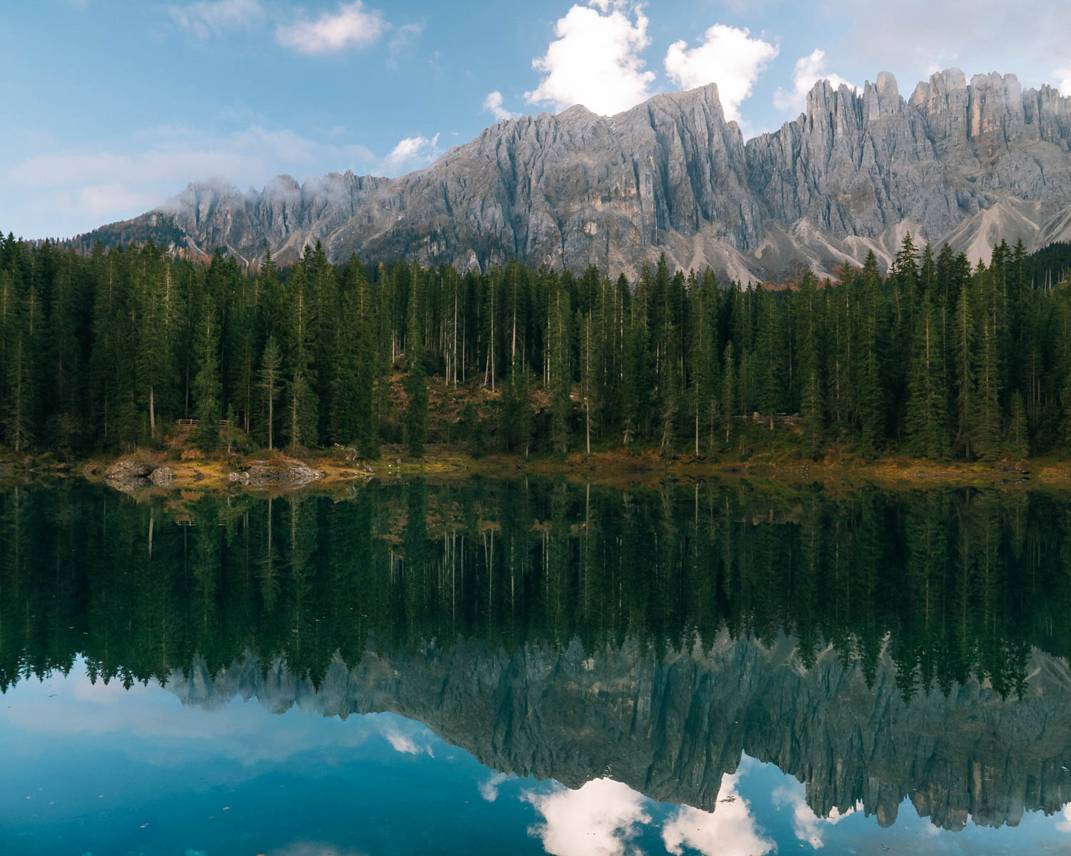 Lago di Carezza reflects the jagged peaks of the Dolomites and the surrounding evergreen forest. The water's surface creates a perfect mirror image of the mountains and sky.