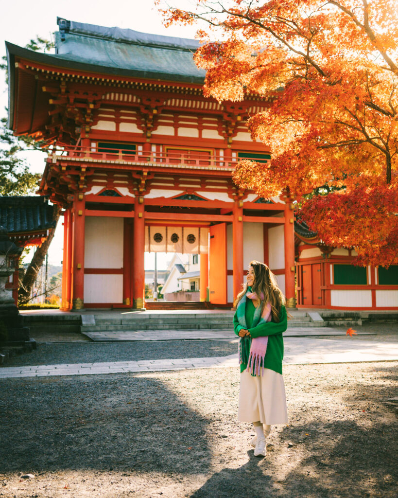 Woman in green sweater and pink scarf walking under autumn leaves at a traditional red temple gate in Kyoto, Japan, with warm afternoon light.